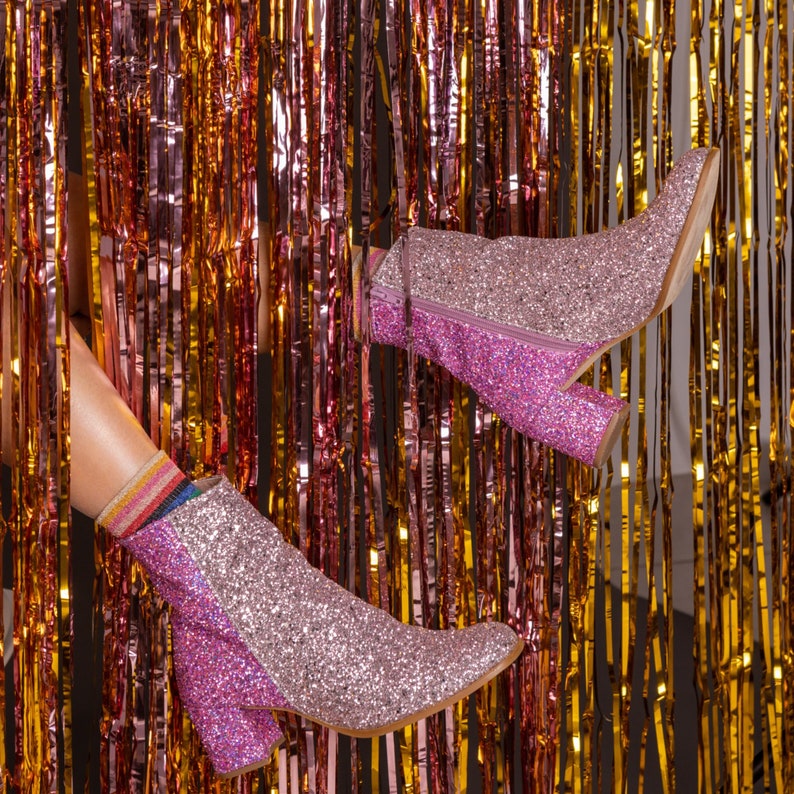 May include: A pair of pink glitter ankle boots with a block heel. The boots are shown against a backdrop of shimmering gold and pink tinsel.