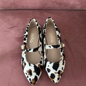 May include: A pair of white, black, and brown animal print flats with a gold buckle. The shoes have a pointed toe and a strap across the top. The brand name "aella" is printed on the inside of each shoe.