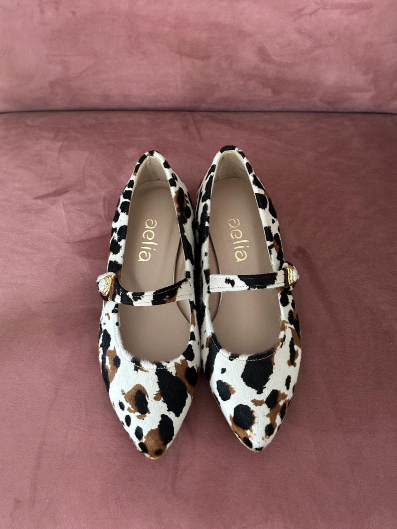 May include: A pair of white, black, and brown animal print flats with a gold buckle. The shoes have a pointed toe and a strap across the top. The brand name "aella" is printed on the inside of each shoe.