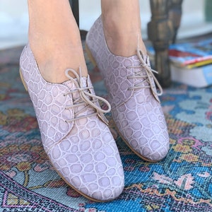Pu&ograve; includere: Un paio di scarpe stringate in pelle color lilla chiaro con un motivo goffrato a coccodrillo. Le scarpe sono su un tappeto a motivi.