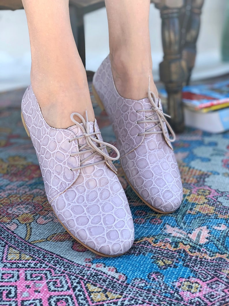 Pu&ograve; includere: Un paio di scarpe stringate in pelle color lilla chiaro con un motivo goffrato a coccodrillo. Le scarpe sono su un tappeto a motivi.