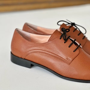 May include: A pair of brown leather lace-up shoes with black laces. The shoes have a slightly rounded toe and a low heel.