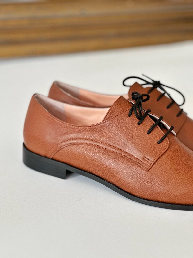 May include: A pair of brown leather lace-up shoes with black laces. The shoes have a slightly rounded toe and a low heel.