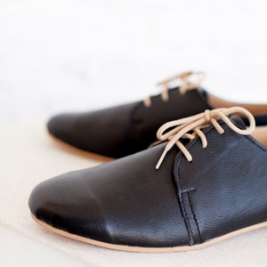 May include: A pair of black leather lace-up shoes with beige laces. The shoes are sitting on a white surface.