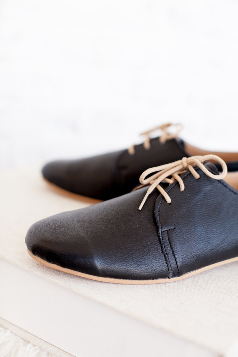 May include: A pair of black leather lace-up shoes with beige laces. The shoes are sitting on a white surface.
