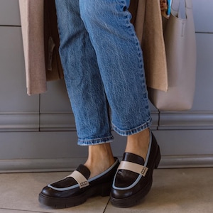 May include: A pair of black leather loafers with a white and blue trim. The loafers have a chunky sole and are worn with blue jeans.
