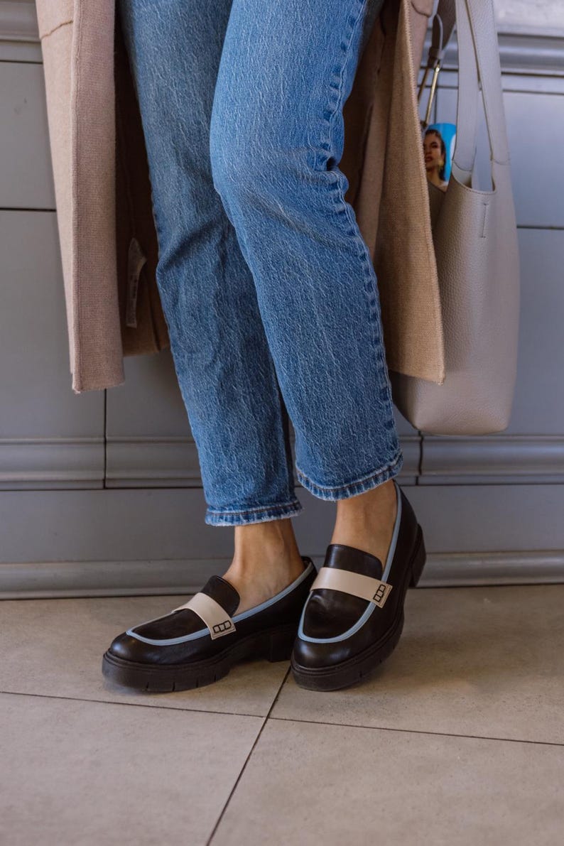 May include: A pair of black leather loafers with a white and blue trim. The loafers have a chunky sole and are worn with blue jeans.