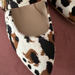 May include: A pair of pointed-toe flats with a brown and white animal print. The shoes have a strap across the top with a gold buckle.