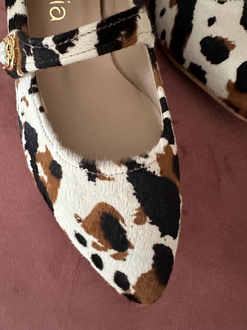 May include: A pair of pointed-toe flats with a brown and white animal print. The shoes have a strap across the top with a gold buckle.
