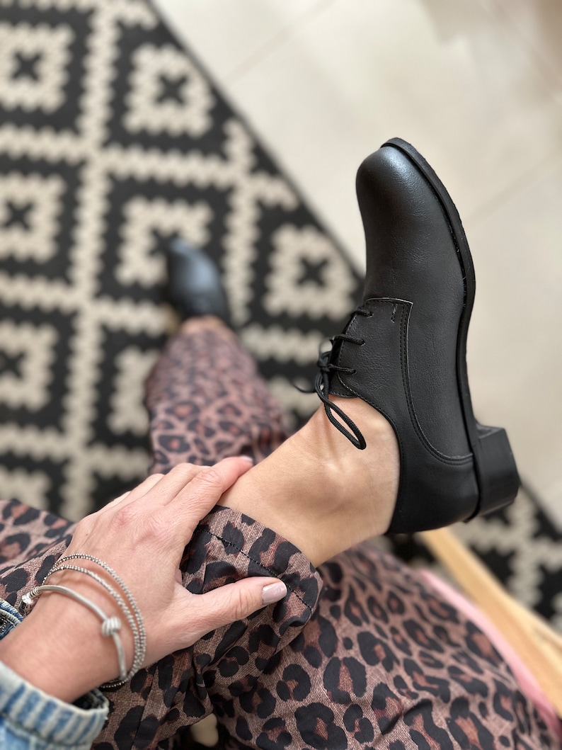 Op de afbeelding: Zwarte leren veterschoenen met een afgeronde neus. De schoenen worden gedragen door iemand die een rok met luipaardprint draagt.
