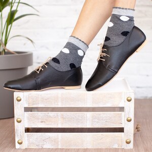 May include: Black leather lace-up shoes with a low heel, worn with grey socks with white polka dots. The shoes are resting on a white wooden crate with brass accents.