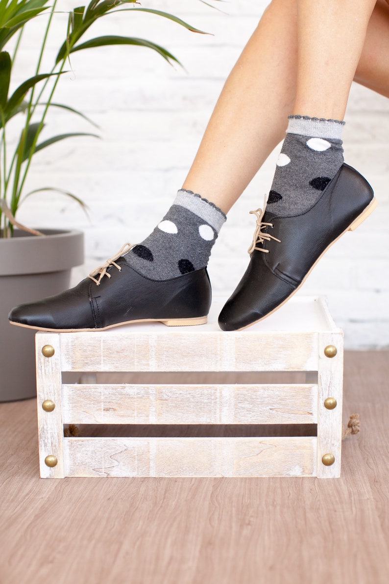 May include: Black leather lace-up shoes with a low heel, worn with grey socks with white polka dots. The shoes are resting on a white wooden crate with brass accents.