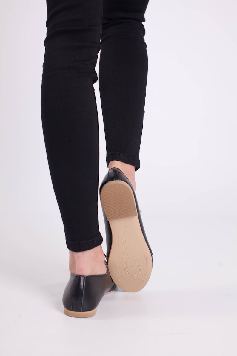 May include: A pair of black leather flats with a pointed toe and a low heel. The shoes are worn by a person wearing black jeans.