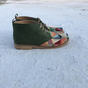 May include: A pair of green suede ankle boots with a colourful geometric pattern on the toe and heel. The boots have a lace-up closure and a brown leather sole.