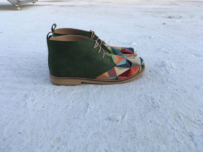 May include: A pair of green suede ankle boots with a colourful geometric pattern on the toe and heel. The boots have a lace-up closure and a brown leather sole.