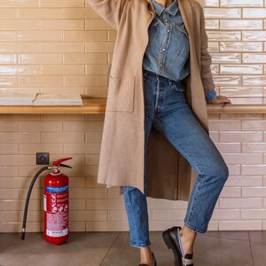 May include: A woman wearing a beige cardigan, a light blue denim shirt, blue jeans, and black loafers with a white stripe. She is wearing a white baseball cap with a red square on the front and sunglasses. She is standing in front of a tiled wall with a red fire extinguisher.