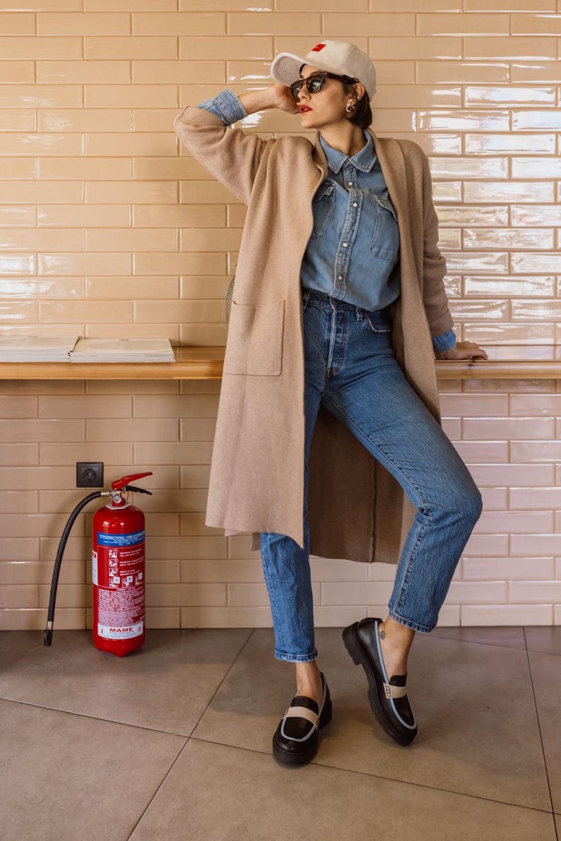 May include: A woman wearing a beige cardigan, a light blue denim shirt, blue jeans, and black loafers with a white stripe. She is wearing a white baseball cap with a red square on the front and sunglasses. She is standing in front of a tiled wall with a red fire extinguisher.