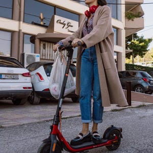 May include: A woman wearing a beige coat, blue jeans, and sunglasses rides a red electric scooter down a city street. She is carrying a white mesh bag.