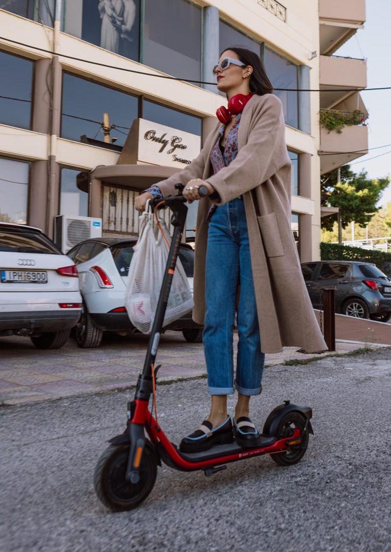 May include: A woman wearing a beige coat, blue jeans, and sunglasses rides a red electric scooter down a city street. She is carrying a white mesh bag.