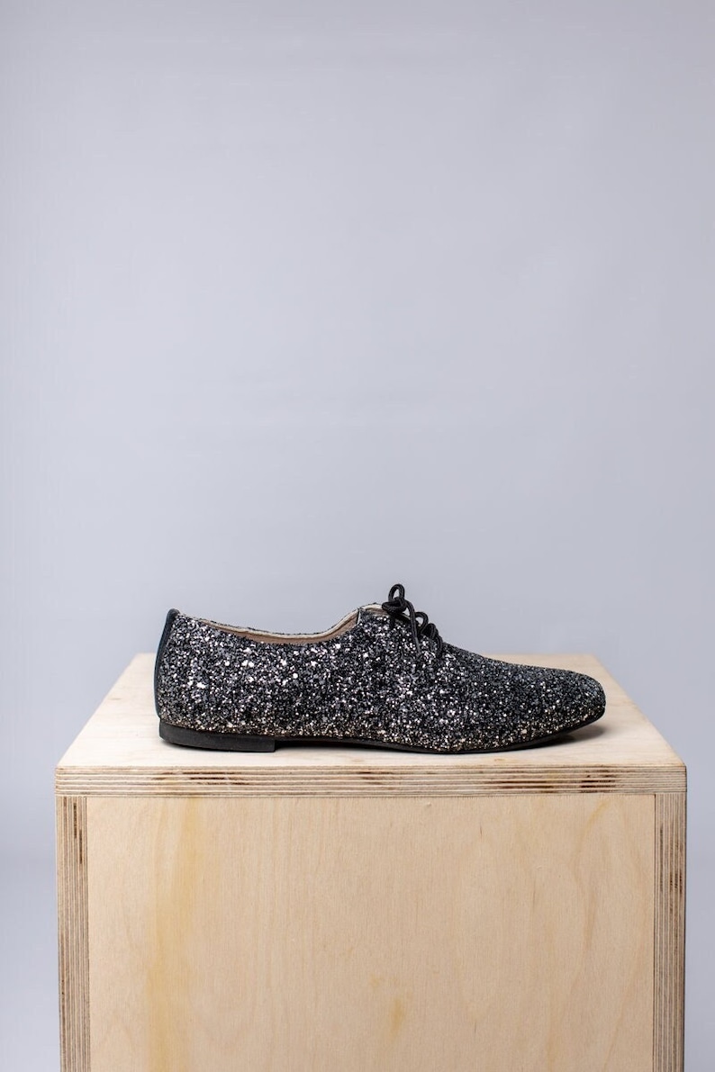 May include: A black and silver glitter flat shoe with a lace-up closure. The shoe is sitting on a wooden surface.