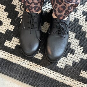 Op de afbeelding: Een paar zwarte leren veterschoenen met een afgeronde neus. De schoenen worden gedragen door iemand die een broek met luipaardprint draagt.