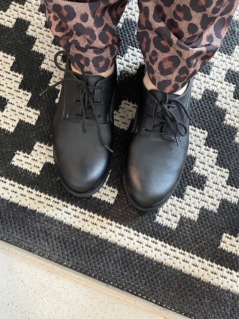 Op de afbeelding: Een paar zwarte leren veterschoenen met een afgeronde neus. De schoenen worden gedragen door iemand die een broek met luipaardprint draagt.