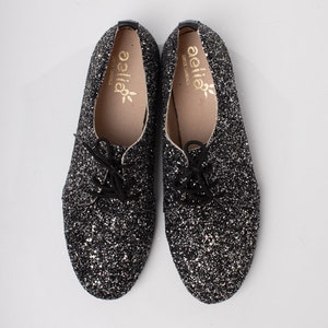 May include: A pair of black glitter lace-up shoes with a gold "aella" logo on the inside of each shoe.