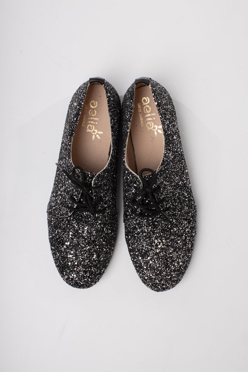 May include: A pair of black glitter lace-up shoes with a gold "aella" logo on the inside of each shoe.