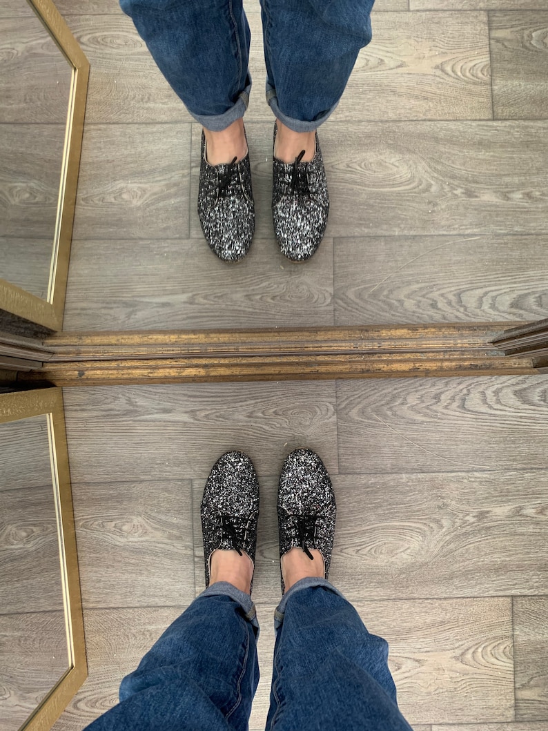 May include: A pair of black and silver glitter shoes with black laces. The shoes are worn by a person wearing blue jeans.
