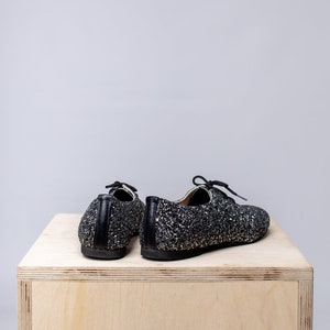 May include: A pair of black sparkly shoes with laces. The shoes are sitting on a wooden surface.