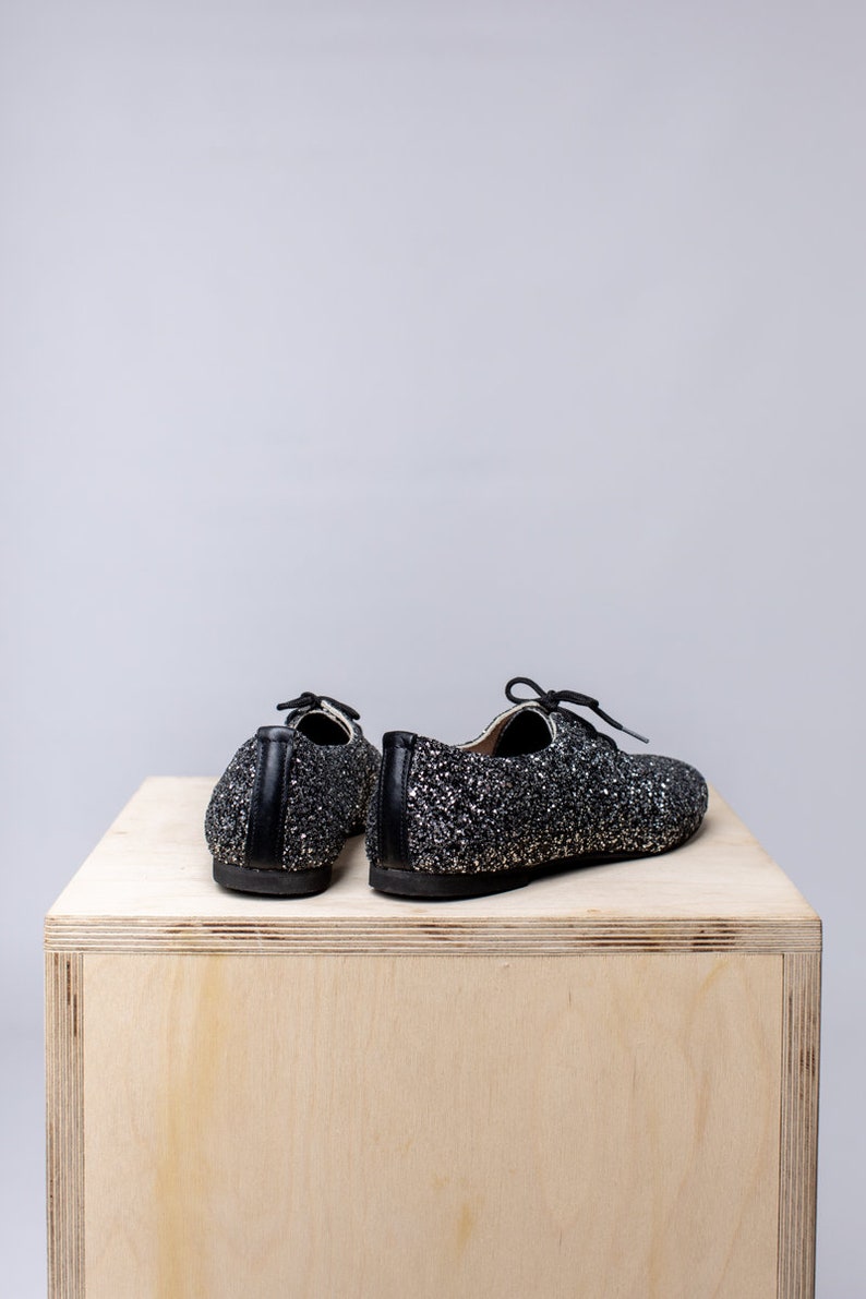 May include: A pair of black sparkly shoes with laces. The shoes are sitting on a wooden surface.