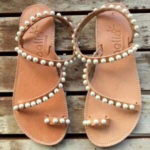 May include: A pair of tan leather sandals with white pearl embellishments. The sandals have a toe loop and an ankle strap. The sandals are made by the brand "Aellia Greek Sandals".