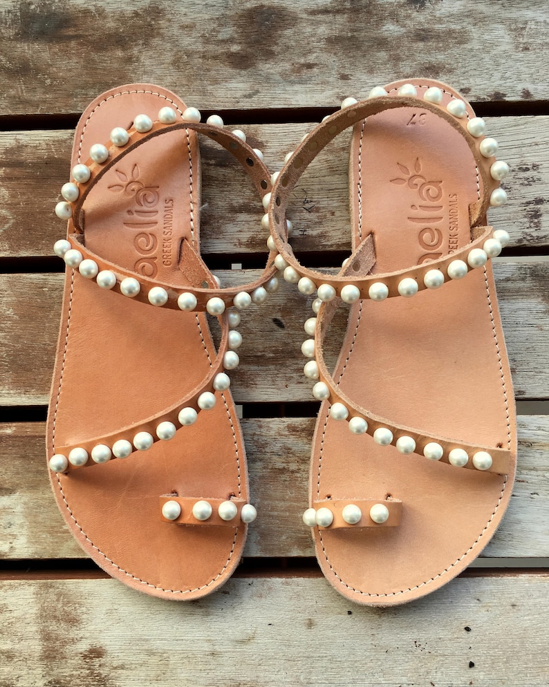 May include: A pair of tan leather sandals with white pearl embellishments. The sandals have a toe loop and an ankle strap. The sandals are made by the brand "Aellia Greek Sandals".