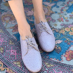 Pu&ograve; includere: Un paio di scarpe con lacci in pelle color lilla chiaro con un motivo testurizzato. Le scarpe sono indossate da una persona in piedi su un tappeto colorato con motivi.