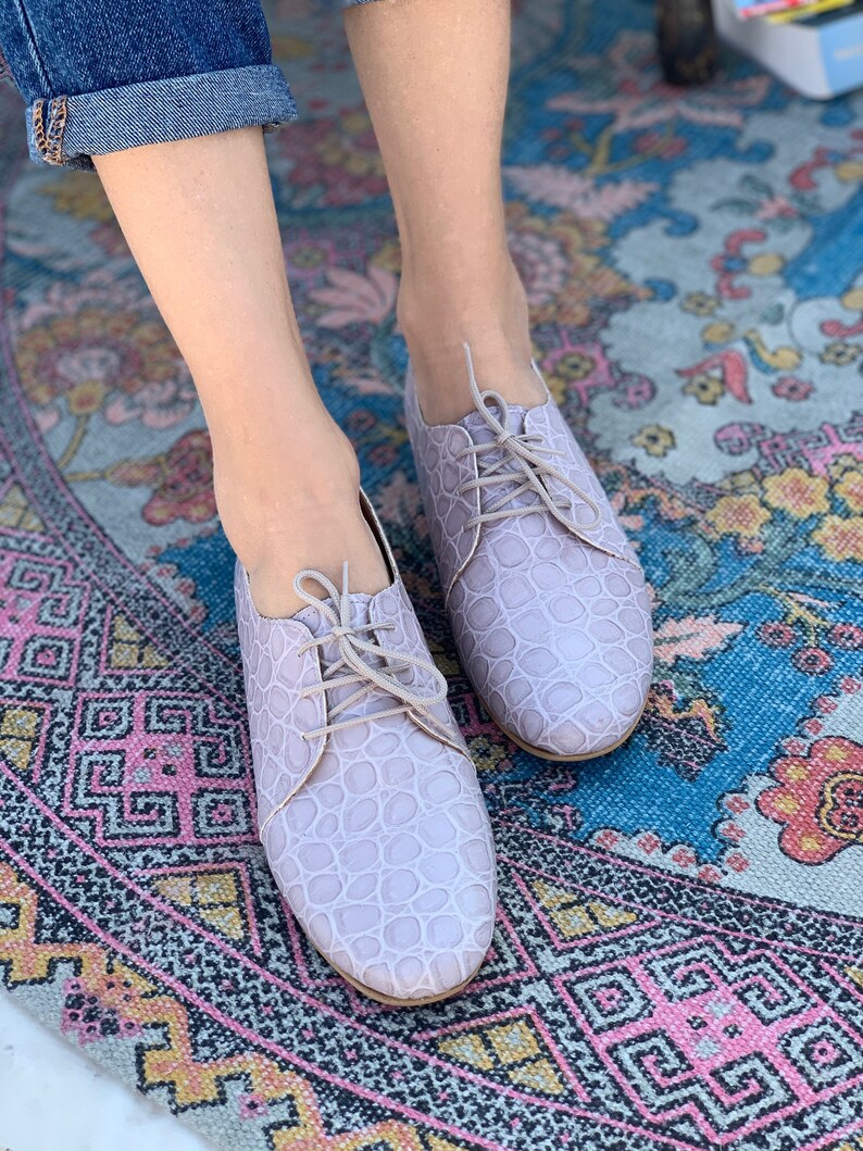 Pu&ograve; includere: Un paio di scarpe con lacci in pelle color lilla chiaro con un motivo testurizzato. Le scarpe sono indossate da una persona in piedi su un tappeto colorato con motivi.