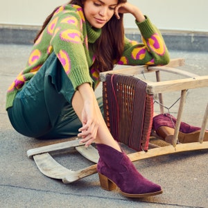 Pu&ograve; includere: Una donna indossa un maglione a motivi verdi e rosa e pantaloni verde scuro &egrave; seduta su una sedia di legno con una seduta intrecciata. Indossa stivali da caviglia in pelle scamosciata color borgogna con punta.
