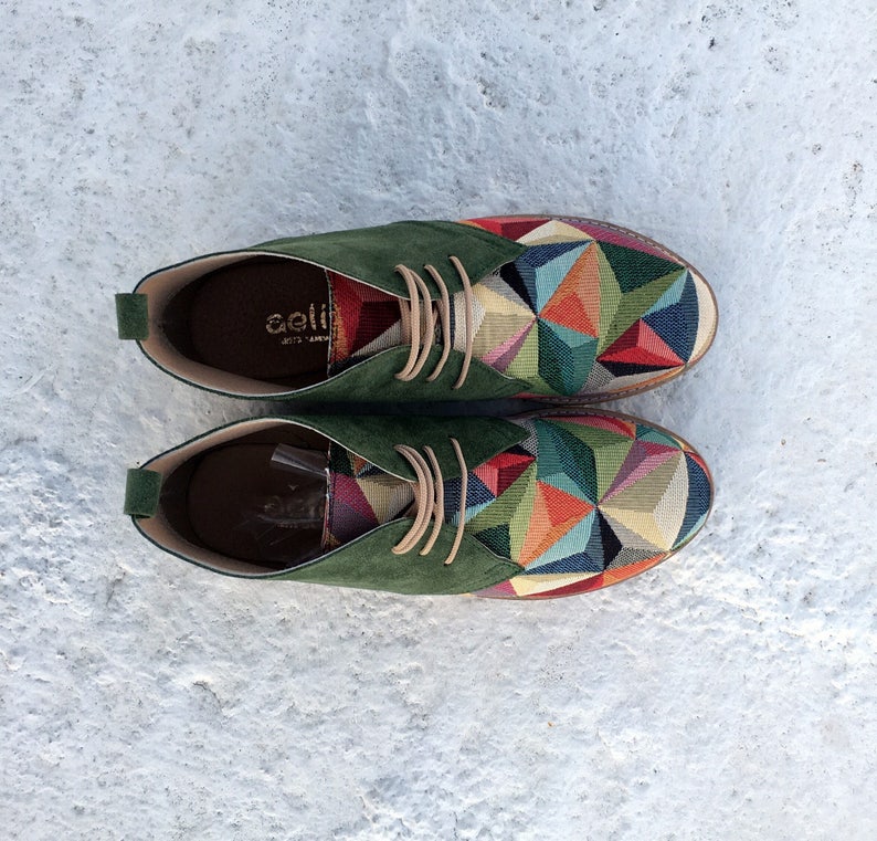 May include: A pair of green suede shoes with a colourful geometric pattern. The shoes have laces and a rounded toe.