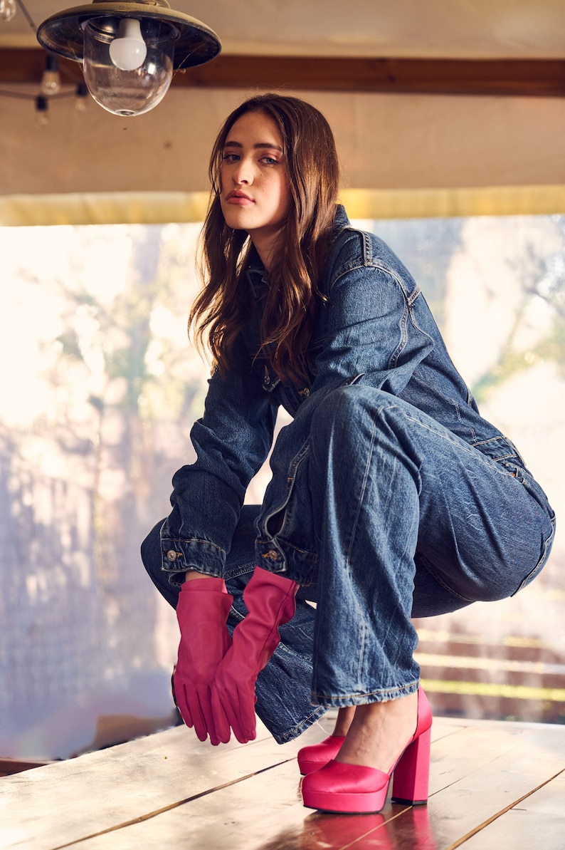 Puede incluir: Una persona con un mono vaquero, guantes rosas y tacones de plataforma rosas. La persona est&aacute; en cuclillas sobre una superficie de madera.