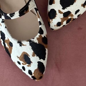 May include: A pair of pointed-toe flats with a cow print pattern. The shoes have a strap across the top with a gold buckle.