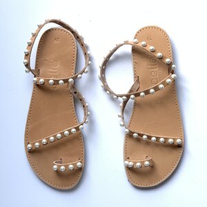May include: A pair of tan leather sandals with white pearl embellishments. The sandals have a toe loop and an ankle strap. The sandals are labeled "aelita" on the footbed.