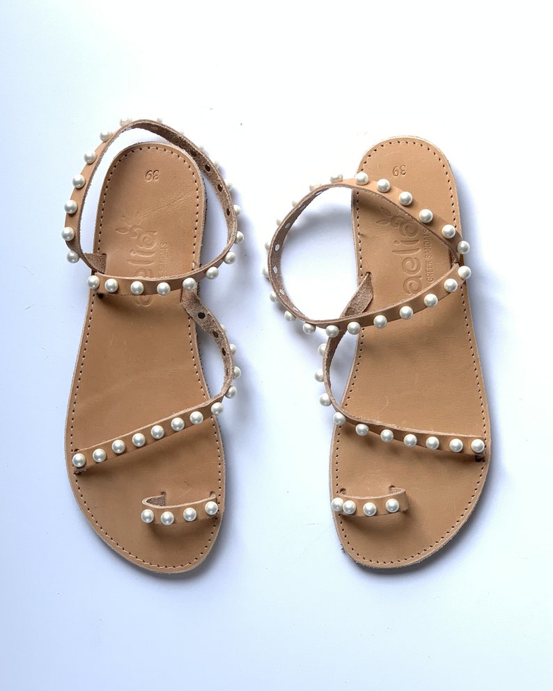 May include: A pair of tan leather sandals with white pearl embellishments. The sandals have a toe loop and an ankle strap. The sandals are labeled "aelita" on the footbed.