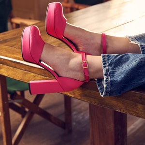 Puede incluir: Un par de tacones de plataforma rosa brillante con correas de tobillo. Los tacones descansan sobre una mesa de madera.
