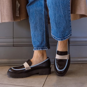 May include: A pair of black leather loafers with a white and light blue trim. The loafers have a chunky heel and a square toe.