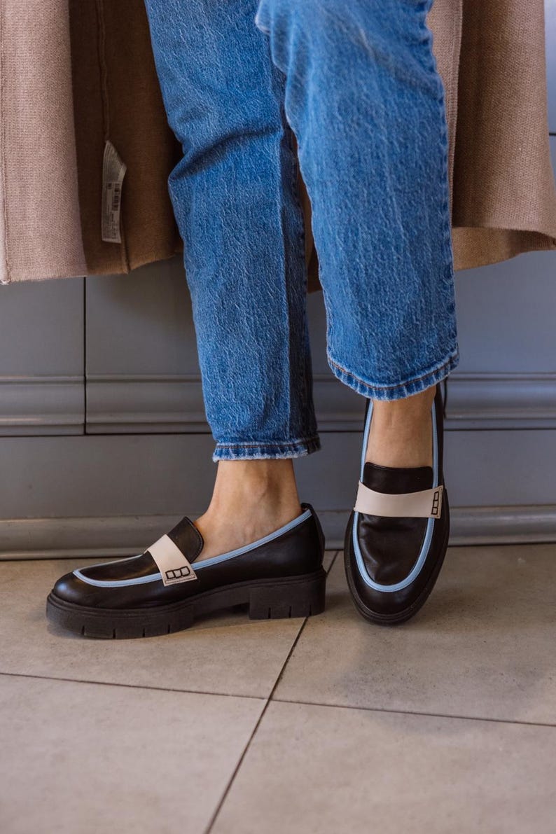 May include: A pair of black leather loafers with a white and light blue trim. The loafers have a chunky heel and a square toe.