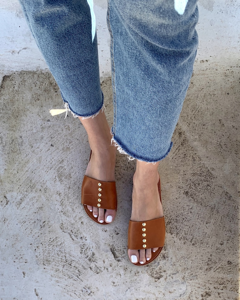 May include: A pair of brown leather sandals with gold studs. The sandals have a simple design and are perfect for summer.