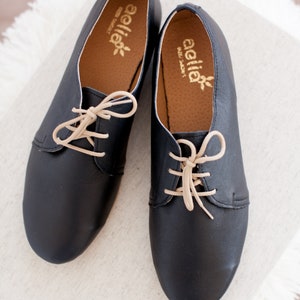 May include: A pair of black leather lace-up shoes with beige laces. The shoes have a rounded toe and a low heel. The brand name "aella" is printed on the inside of each shoe.