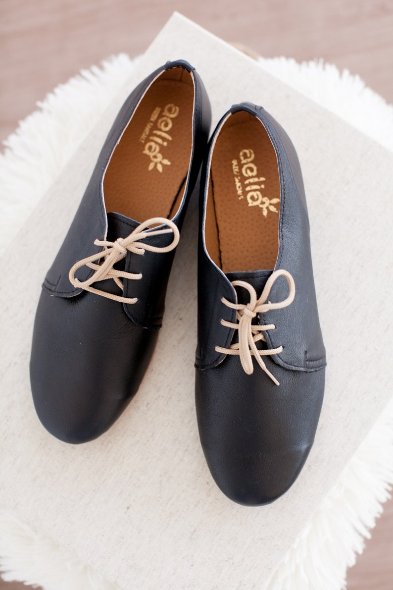 May include: A pair of black leather lace-up shoes with beige laces. The shoes have a rounded toe and a low heel. The brand name "aella" is printed on the inside of each shoe.