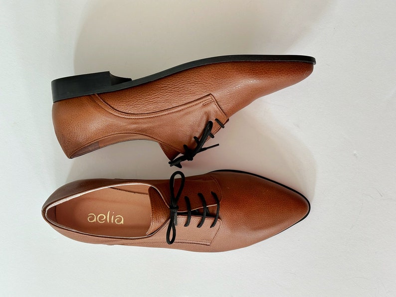 May include: A pair of brown leather lace-up shoes with black laces. The shoes have a pointed toe and a low heel. The brand name "aelia" is printed on the inside of one shoe.