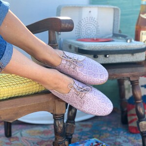Pu&ograve; includere: Un paio di scarpe con lacci in pelle color lavanda con un motivo testurizzato. Le scarpe sono indossate da una persona seduta su una sedia di legno.