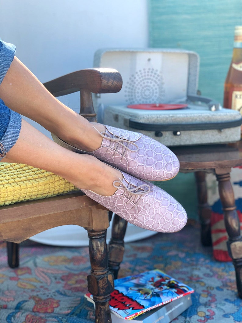 Pu&ograve; includere: Un paio di scarpe con lacci in pelle color lavanda con un motivo testurizzato. Le scarpe sono indossate da una persona seduta su una sedia di legno.